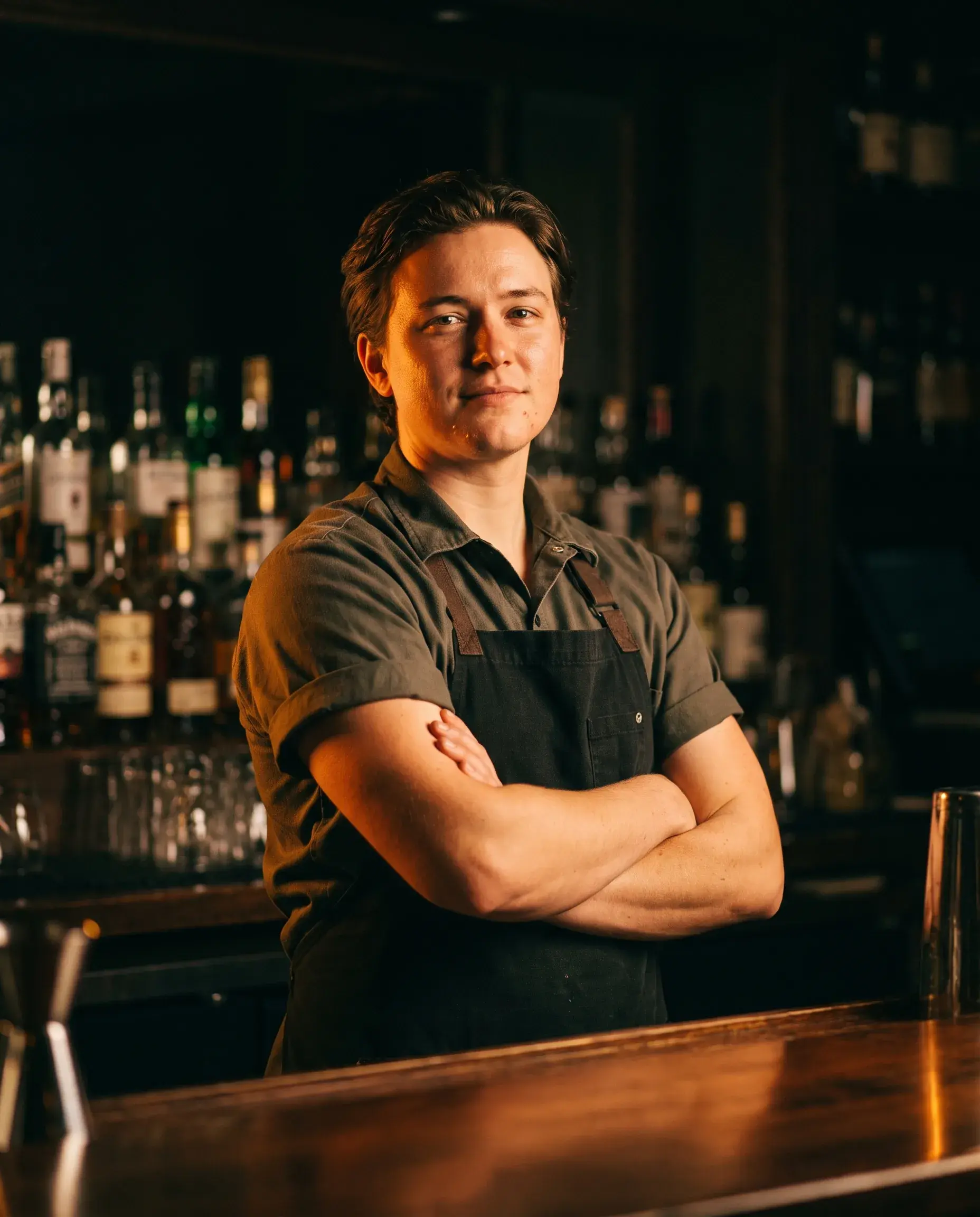 A bartender standing behind a dark bar, arms crossed